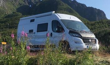 Ein weißer Campingvan steht vor grünen Bergen und blühenden rosa Wildblumen