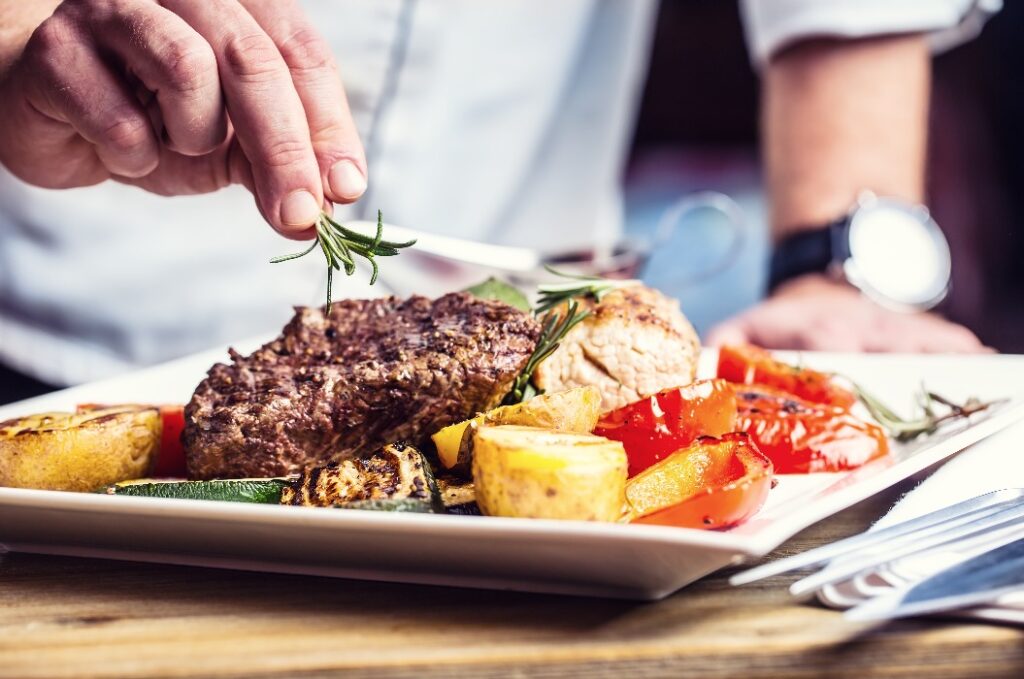 Koch in einer Hotel- oder Restaurantküche, der ausschließlich mit den Händen arbeitet. Zubereitetes Rindersteak mit Gemüsebeilage.