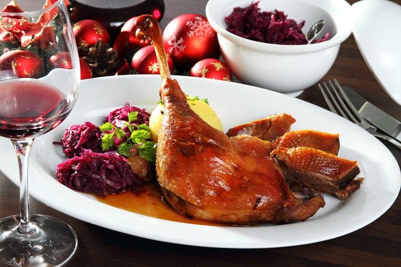 Auf einem weißen Teller liegt knusprige Gänsekeule mit Rotkohl und Kloß neben Rotwein
